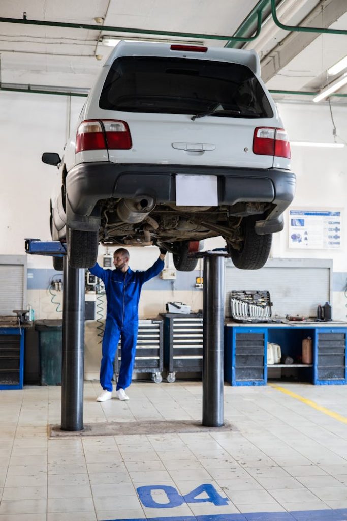 Mechanic in blue coverall inspecting a lifted car in an auto repair shop.